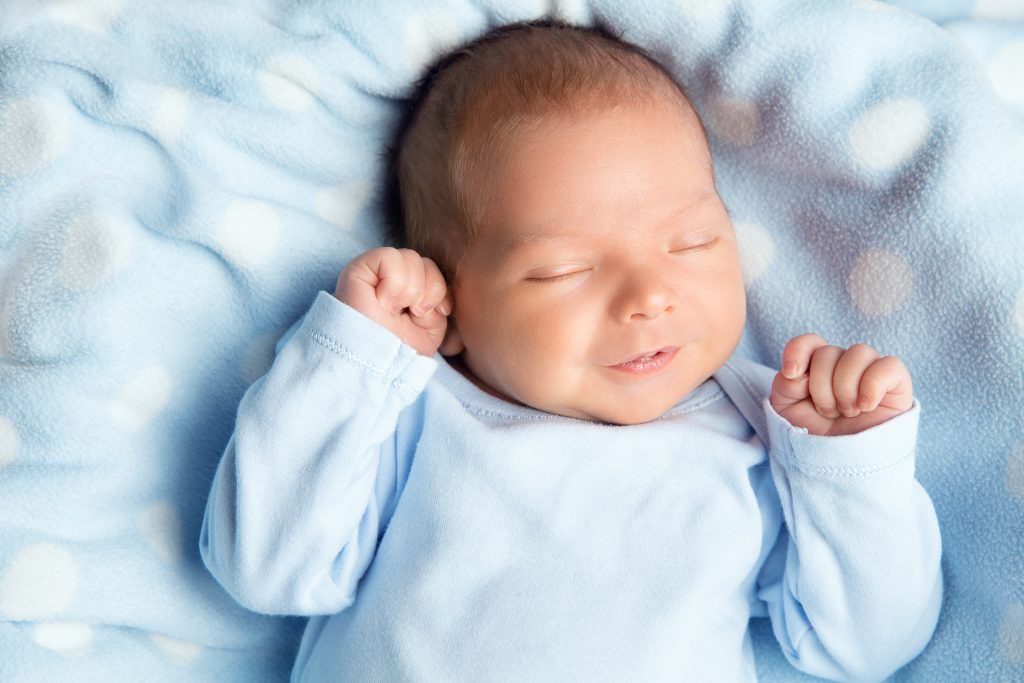 Newborn Baby Sleeping Smiling. Cute Infant Child in Wrap Bodysuit. New Born Little Boy smile in Blue Clothes lying on Soft Blanket. Small Kid Face Close up Portrait holding Hands First