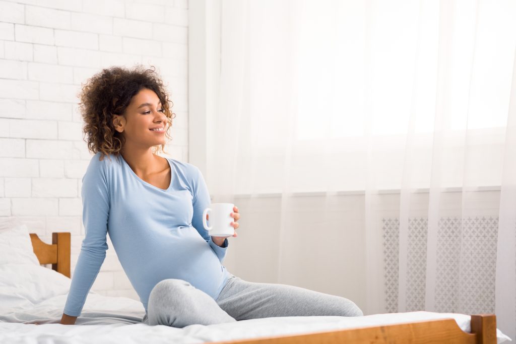 Relaxed pregnant woman drinking tea on bed at home and looking at free space, panorama