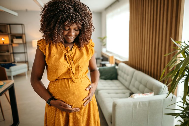 A pregnant woman with dark hair and dark skin cradles her belly. She is standing next to a couch.