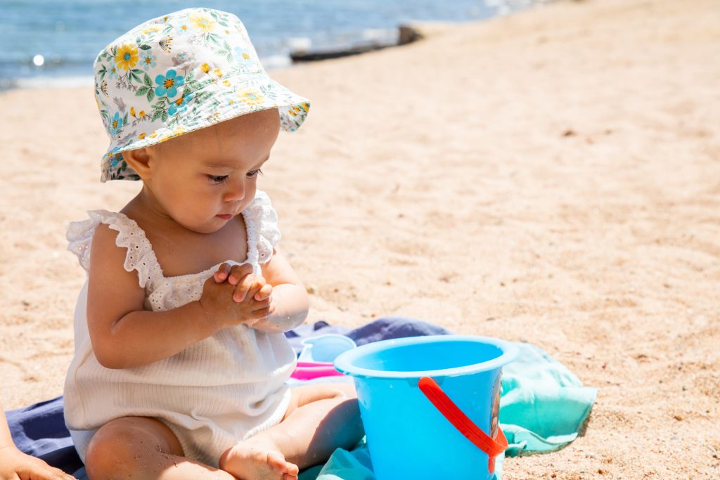 Closeup of baby in sun hat sitting on a beach by the sea playing with blue bucket toy