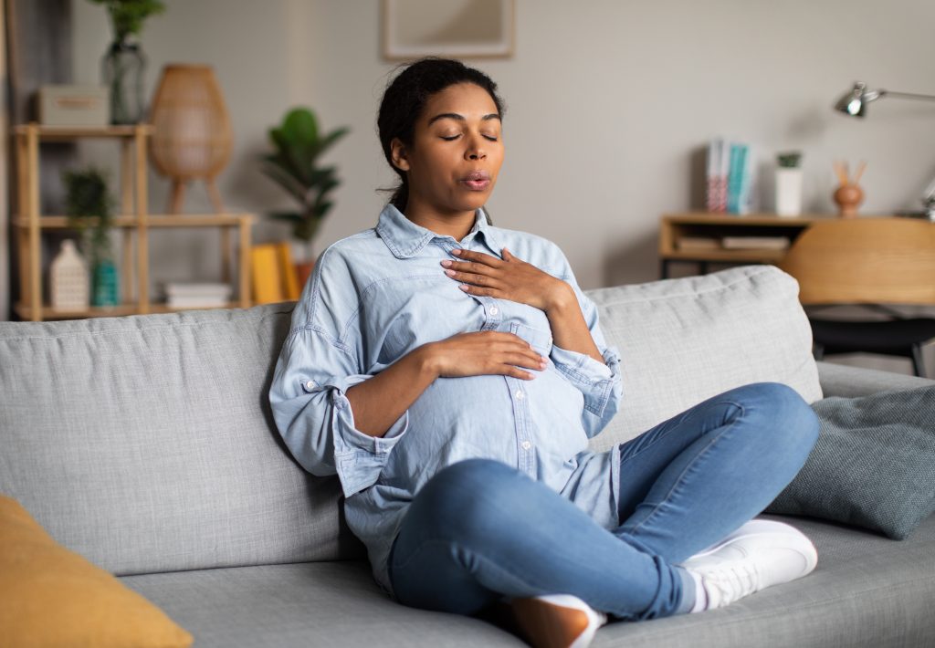 Pregnant woman doing breathing exercise with eyes closed relaxing sitting on couch at home